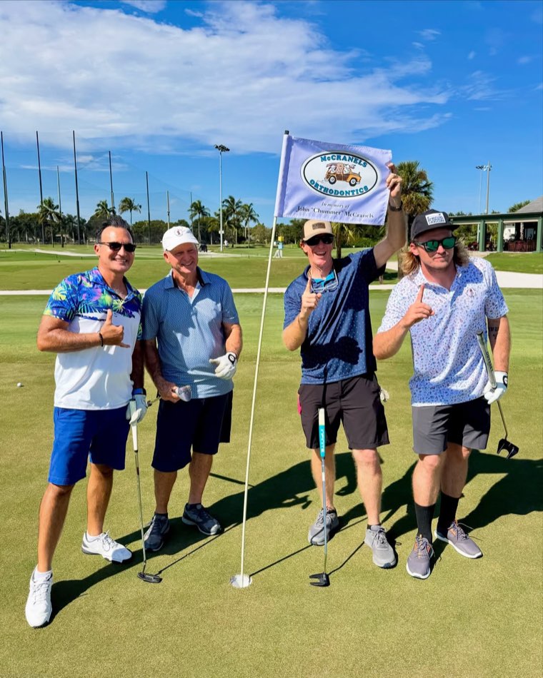 A foursome celebrates on the green at the Surfing Florida Museum Charity Golf Tournament, holding a McCranels Orthodontics flag in memory of John "Chummer" McCranels